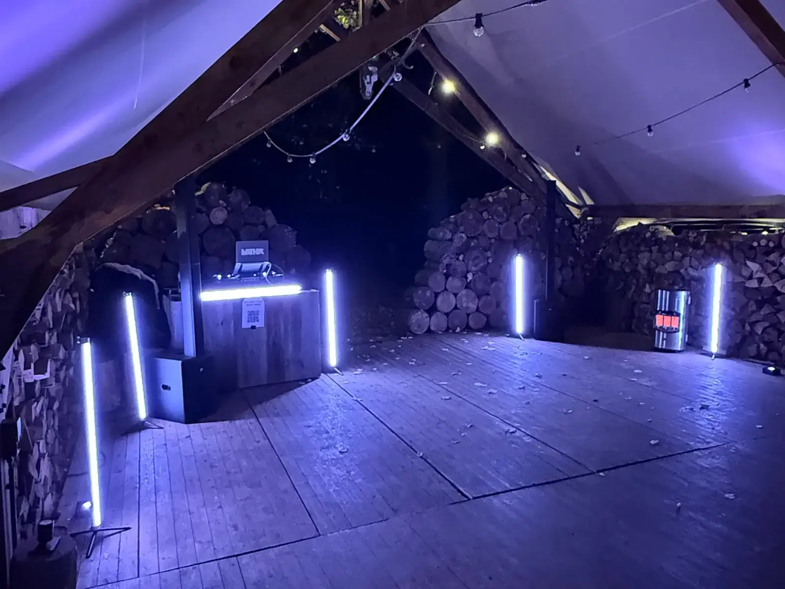 DJ-booth met paarse lichtbuizen tussen houtstapels onder een tentdak