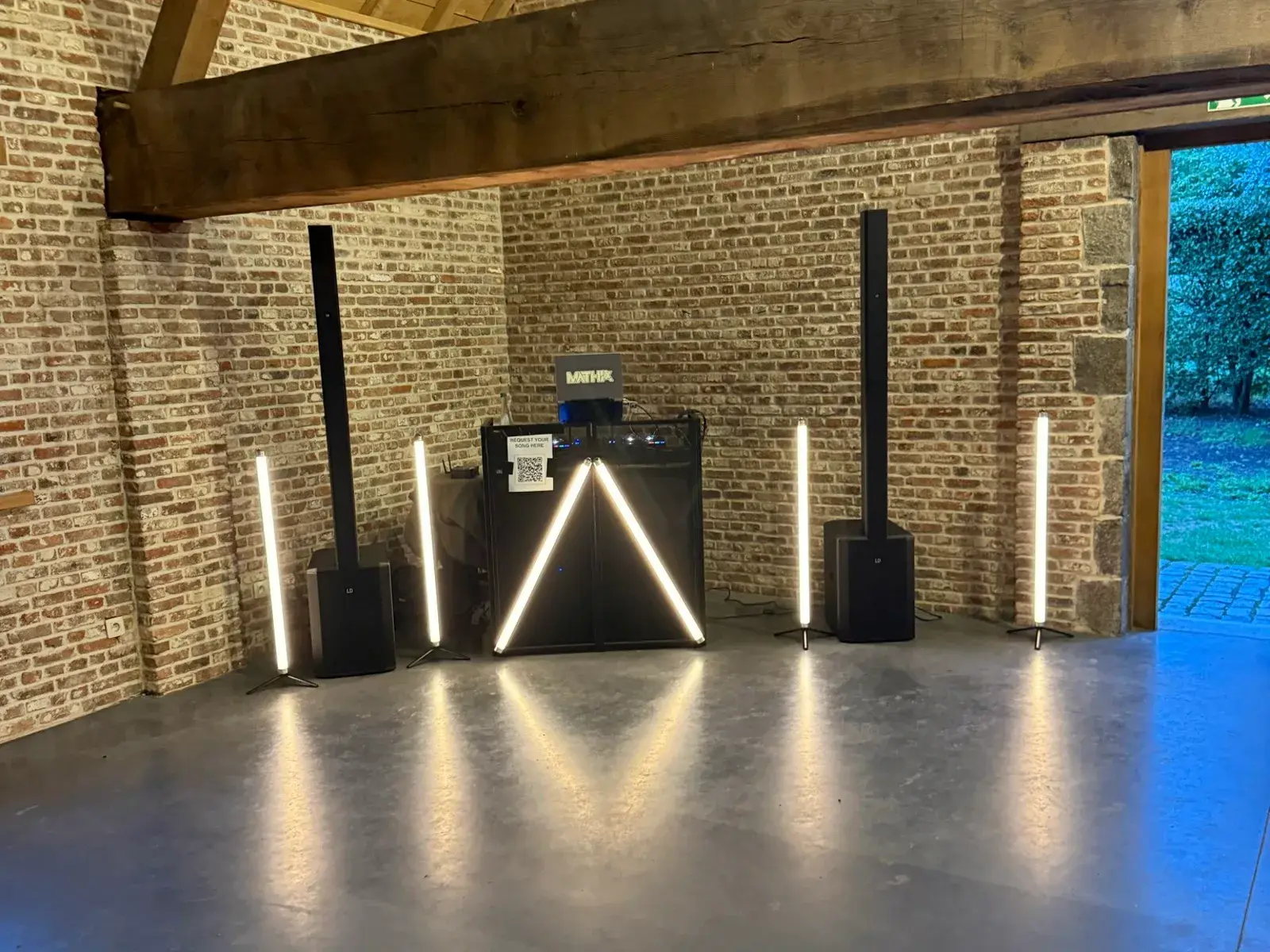 DJ-set met warme lichtbuizen onder houten balken in een bakstenen zaal, klaar voor het feest